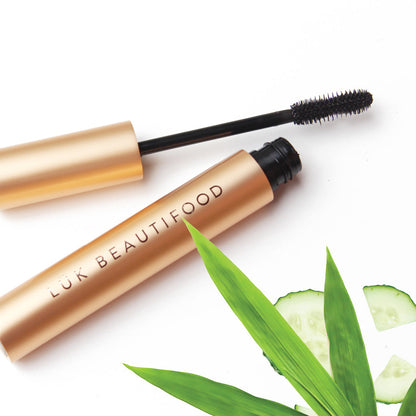 Lük Beautifood Lash Nourish natural mascara, gold tube with the wand lifted, beside fresh cucumber and bamboo leaves